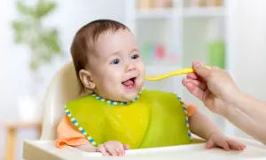 Uśmiechnięte niemowlę w śliniaczku czeka na kolejną łyżeczkę. Rodzic zastanawia się, ile słoiczków dziennie dla niemowląt to optymalna ilość.