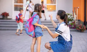 Mama żegna córkę idącą do zerówki. Ile lat ma się w zerówce? Zazwyczaj 6. Dzieci z plecakami wchodzą do szkoły.