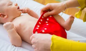 Mama pokazuje, jak zakładać pieluchy tetrowe. Dziecko leży na białym przewijaku, a mama zapina czerwoną pieluszkę na zatrzaski.