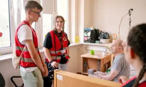 Młodzi wolontariusze PCK rozmawiają z seniorem w pokoju. Zastanawiasz się, ile kosztuje opiekunka z PCK?