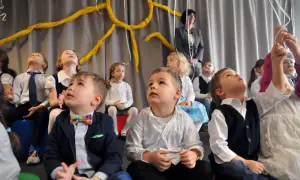 Dzieci w odświętnych strojach, jak na pasowanie przedszkolaka, patrzą w górę. Chłopiec w garniturku i kolorowej muszce.