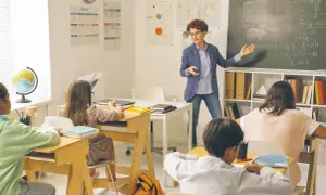Nauczycielka tłumaczy zadanie na tablicy. Dzieci siedzą w ławkach. Ile godzin pracuje pomoc nauczyciela w przedszkolu?
