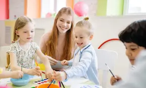 Uśmiechnięta opiekunka pomaga dzieciom malować. Zastanawiasz się, ile zarabia opiekun w przedszkolu?