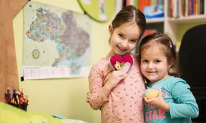 Dwie uśmiechnięte dziewczynki trzymają serduszka. W tle mapa i półki z książkami. Ile godzin w przedszkolu jest bezpłatnych?