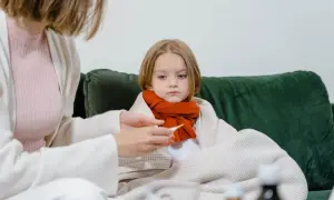 Smutna dziewczynka owinięta kocem, z szalikiem na szyi, czeka na powrót do zdrowia po grypie. Mama mierzy jej temperaturę.