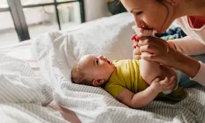 Mama całuje stópki niemowlaka, który leży na łóżku. Widać, że dziecko szybko rośnie, kiedy podwojenie masy urodzeniowej jest już za nim.