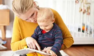 Mama uczy dziecko mówić, czytając mu książeczkę. W tle widać łóżeczko dziecięce.