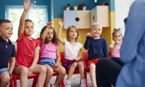 Dzieci na czerwonych krzesłach podnoszą ręce, odpowiadając na pytanie o to, ile lat się chodzi do przedszkola.