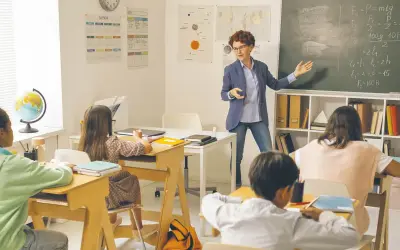 Nauczycielka tłumaczy zadanie na tablicy. Dzieci siedzą w ławkach. Ile godzin pracuje pomoc nauczyciela w przedszkolu?