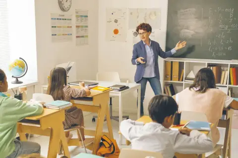 Nauczycielka tłumaczy zadanie na tablicy. Dzieci siedzą w ławkach. Ile godzin pracuje pomoc nauczyciela w przedszkolu?