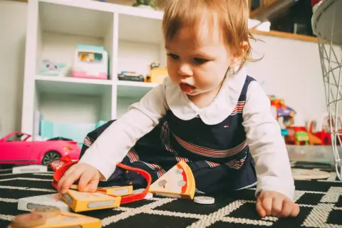 Małe dziecko bawi się zabawkami na dywanie. W tle regał z zabawkami. Żłobek od jakiego wieku? Od 6 miesiąca życia.