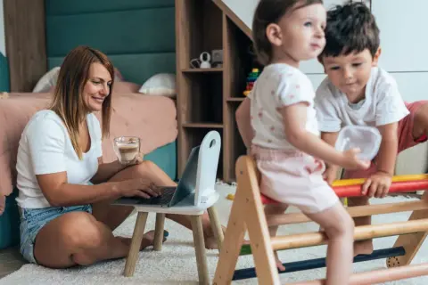 Kobieta pracuje na laptopie, pijąc kawę, podczas gdy dzieci bawią się obok. Pokazuje, jak pogodzić pracę z opieką nad dzieckiem.