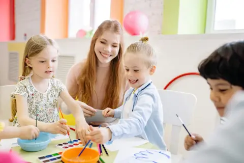 Uśmiechnięta opiekunka pomaga dzieciom malować. Zastanawiasz się, ile zarabia opiekun w przedszkolu?