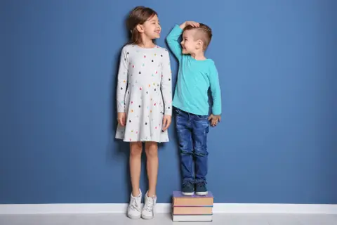 Chłopiec mierzy wzrost dziewczynki, która stoi na książkach. Zastanawiają się, ile cm rośnie dziecko w ciągu roku.