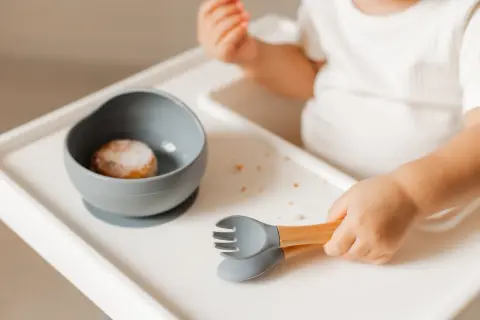 Małe rączki chwytają sztućce, gdy dziecko samo je. Miseczka z jedzeniem i okruszki na tacce.