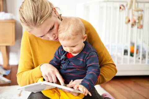 Mama uczy dziecko mówić, czytając mu książeczkę. W tle widać łóżeczko dziecięce.