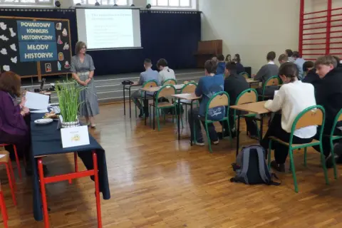 Uczniowie w sali lekcyjnej słuchają nauczycielki podczas konkursu historycznego. Jak wygrać z nauczycielem? Wiedzą, że kluczem jest wiedza!