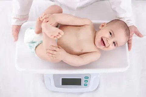 Uśmiechnięte niemowlę na wadze, pokazuje, jak szybko rośnie niemowlę. Rączki i nóżki w górze, radość na twarzy.