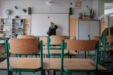 Klasa szkolna z pustymi ławkami. Nauczycielka pisze na tablicy. Zastanawiasz się, ile zarabia pomoc nauczyciela na pół etatu?