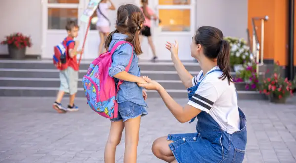 Mama żegna córkę idącą do zerówki. Ile lat ma się w zerówce? Zazwyczaj 6. Dzieci z plecakami wchodzą do szkoły.