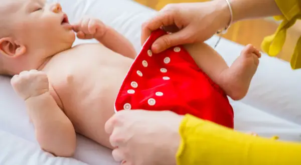 Mama pokazuje, jak zakładać pieluchy tetrowe. Dziecko leży na białym przewijaku, a mama zapina czerwoną pieluszkę na zatrzaski.