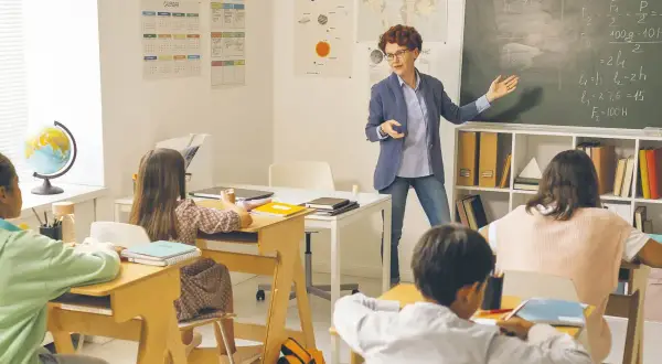 Nauczycielka tłumaczy zadanie na tablicy. Dzieci siedzą w ławkach. Ile godzin pracuje pomoc nauczyciela w przedszkolu?