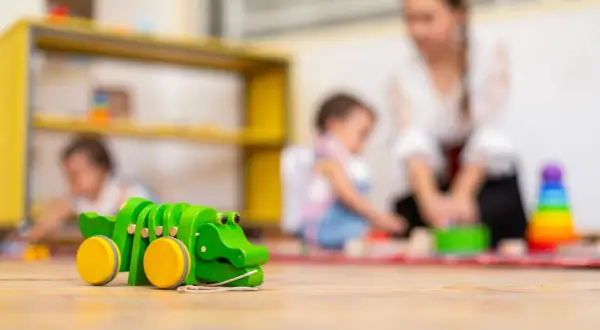Drewniany krokodyl na podłodze żłobka. W tle opiekunka z dziećmi. Ciekawe, ile zarabia opiekun w żłobku państwowym za taką pracę.