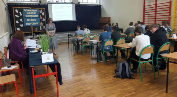 Uczniowie w sali lekcyjnej słuchają nauczycielki podczas konkursu historycznego. Jak wygrać z nauczycielem? Wiedzą, że kluczem jest wiedza!