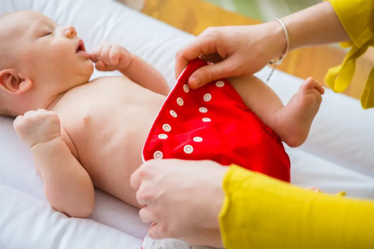 Mama pokazuje, jak zakładać pieluchy tetrowe. Dziecko leży na białym przewijaku, a mama zapina czerwoną pieluszkę na zatrzaski.