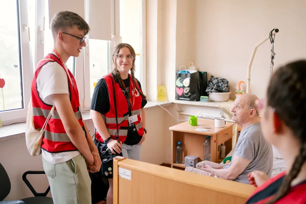 Młodzi wolontariusze PCK rozmawiają z seniorem w pokoju. Zastanawiasz się, ile kosztuje opiekunka z PCK?