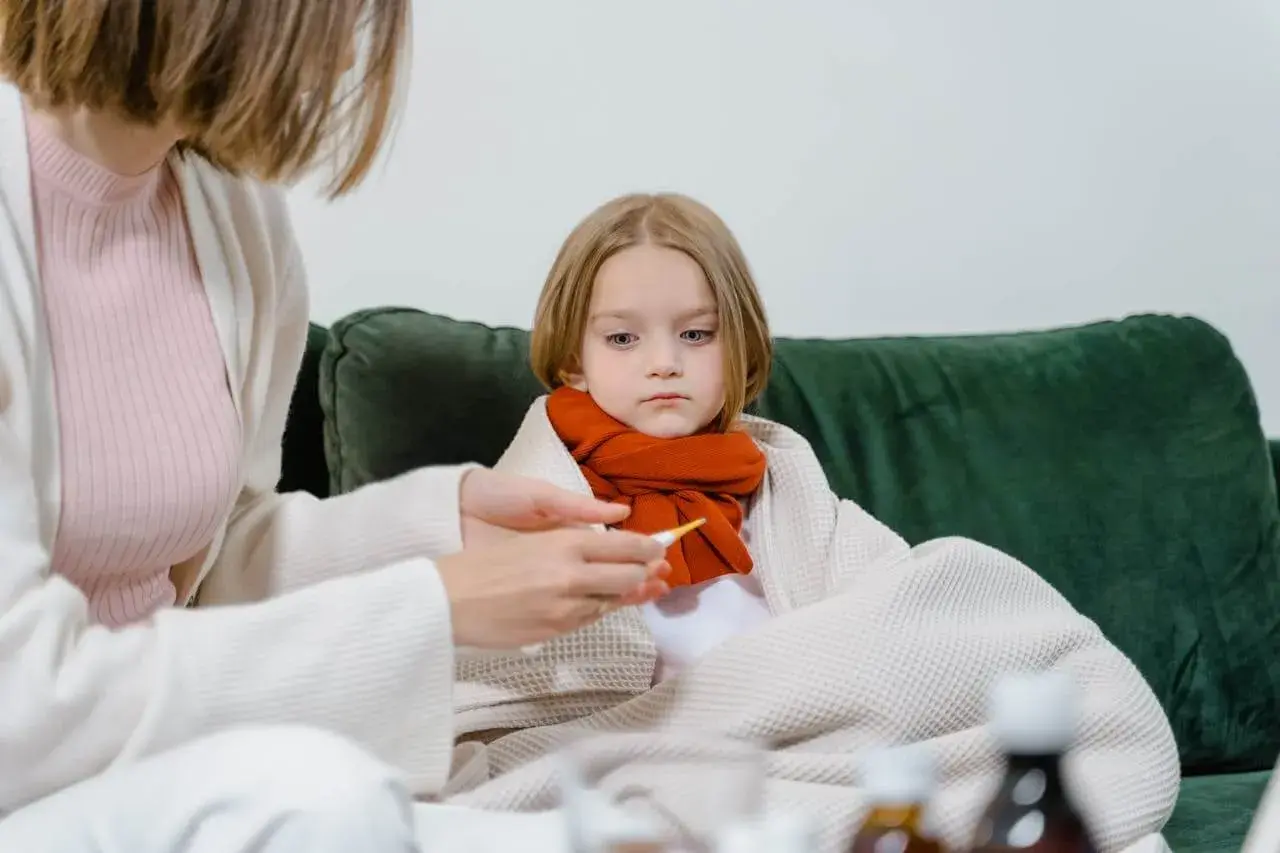 Smutna dziewczynka owinięta kocem, z szalikiem na szyi, czeka na powrót do zdrowia po grypie. Mama mierzy jej temperaturę.
