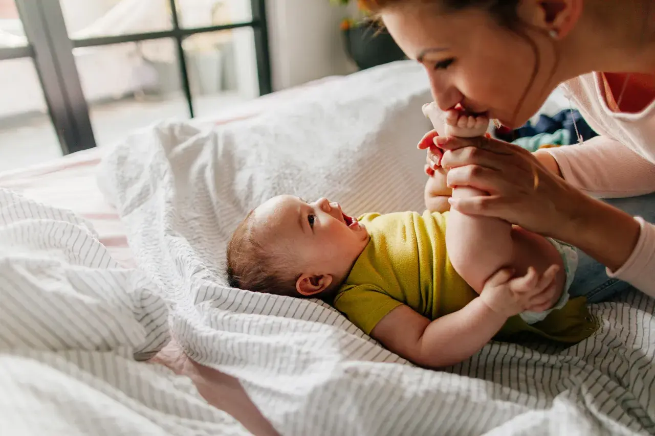 Mama całuje stópki niemowlaka, który leży na łóżku. Widać, że dziecko szybko rośnie, kiedy podwojenie masy urodzeniowej jest już za nim.