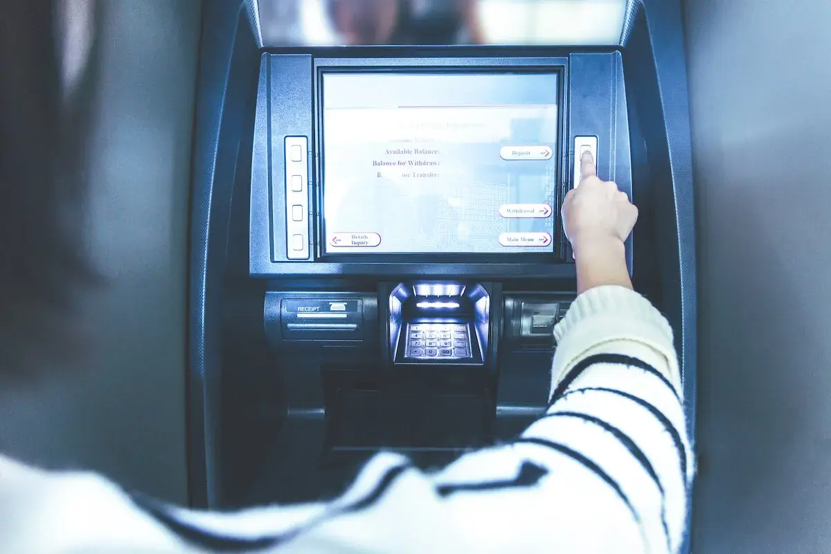 Ręka naciska przycisk na bankomacie. Ekran pokazuje opcje: "Deposit", "Withdrawal", "Main Menu". Czy z konta dziecka można robić przelewy?