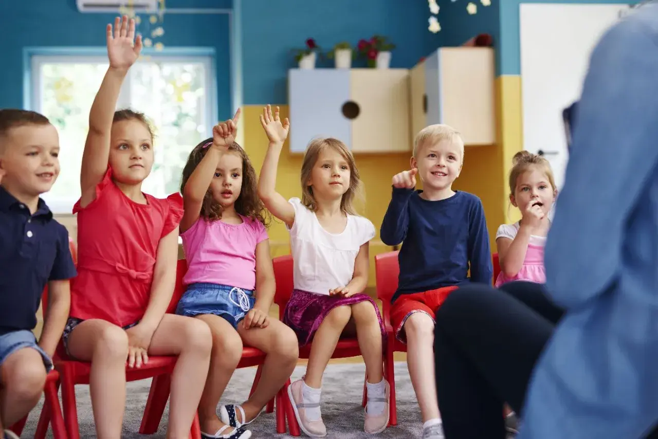 Dzieci na czerwonych krzesłach podnoszą ręce, odpowiadając na pytanie o to, ile lat się chodzi do przedszkola.