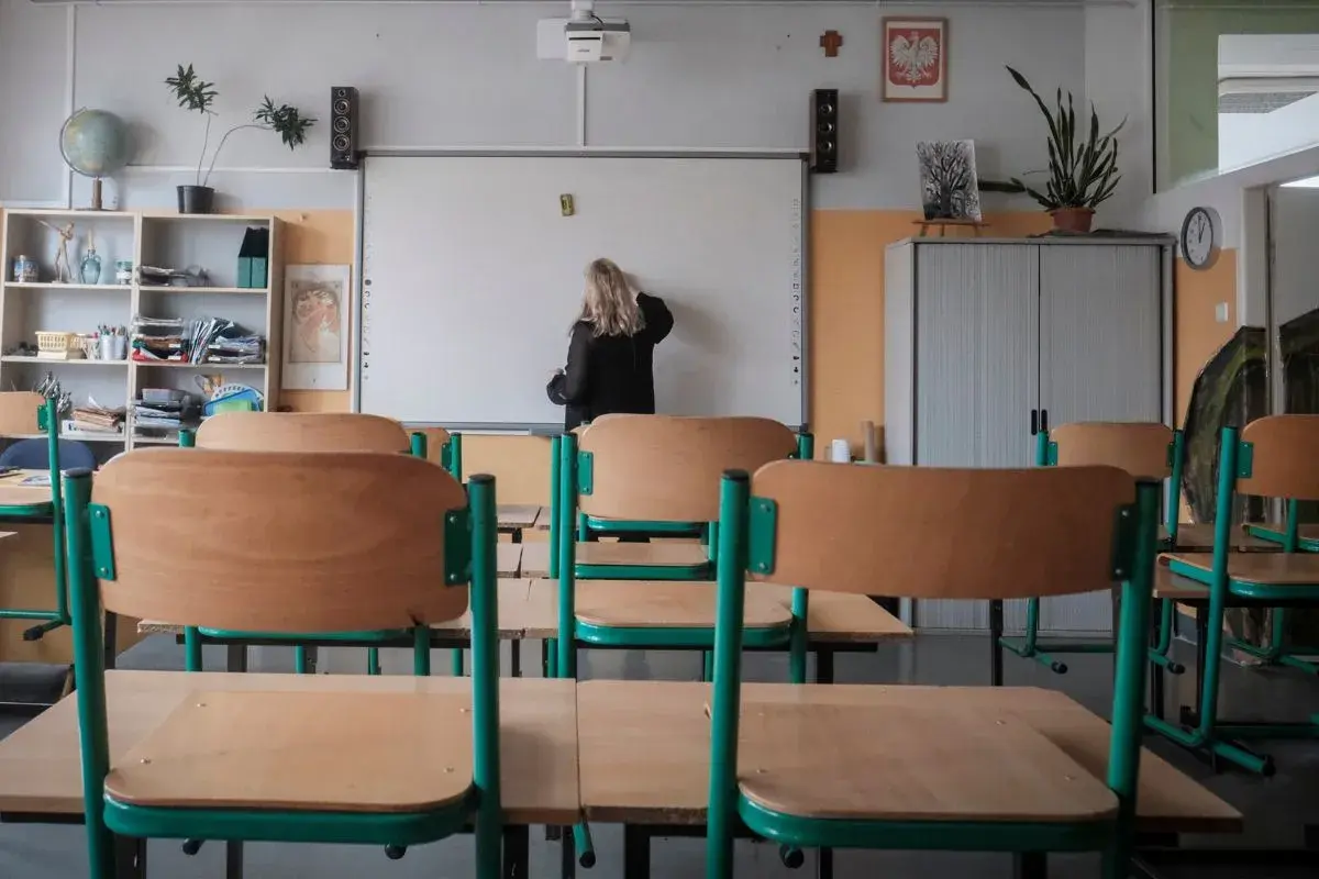 Klasa szkolna z pustymi ławkami. Nauczycielka pisze na tablicy. Zastanawiasz się, ile zarabia pomoc nauczyciela na pół etatu?