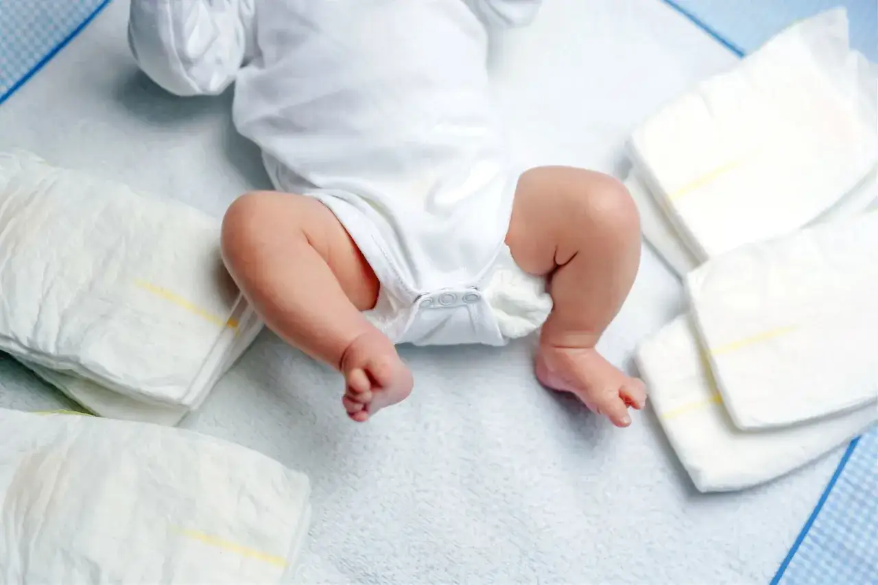Nóżki niemowlaka w białym body, obok stosy pieluszek. Kiedy zmieniać pieluszkę przed czy po karmieniu? To pytanie wielu rodziców.