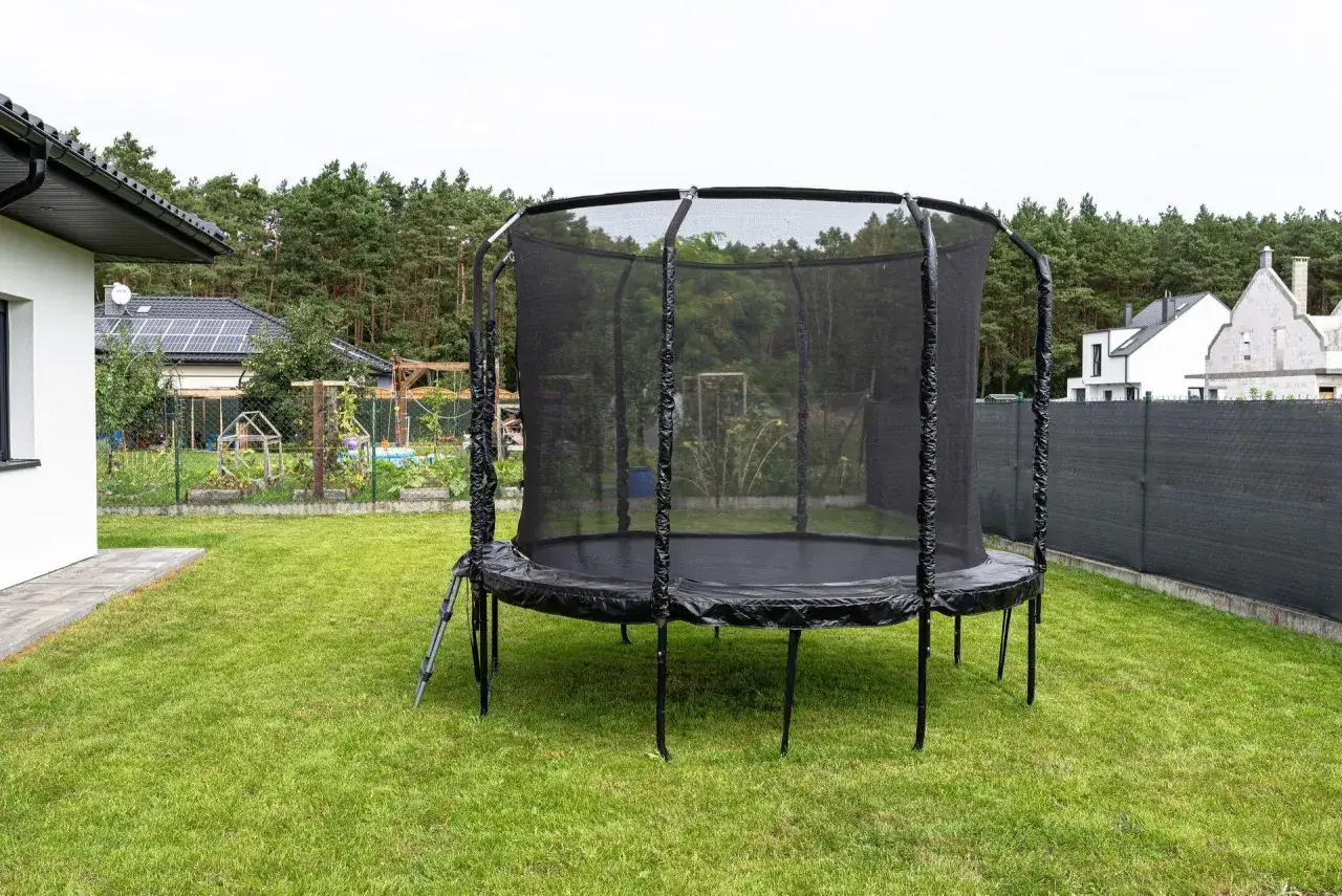 Duża, czarna trampolina stoi na zielonym trawniku. Czy trampolina jest zdrowa? To świetna zabawa i aktywność fizyczna dla całej rodziny.
