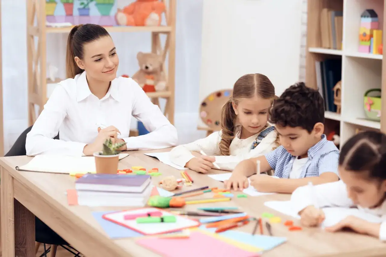 Nauczycielka w przedszkolu prywatnym z uśmiechem obserwuje dzieci rysujące przy stole. Ile godzin pracuje nauczyciel w przedszkolu prywatnym? To zależy od wielu czynników.