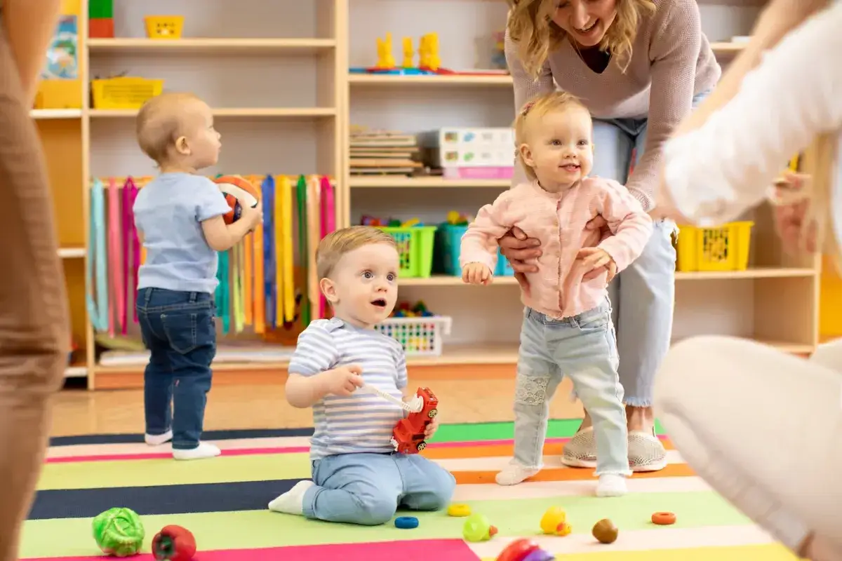 Dzieci bawią się w żłobku. Opiekunka pomaga maluchowi wstać. Ciekawe, ile zarabia pomoc w żłobku za taką pracę.