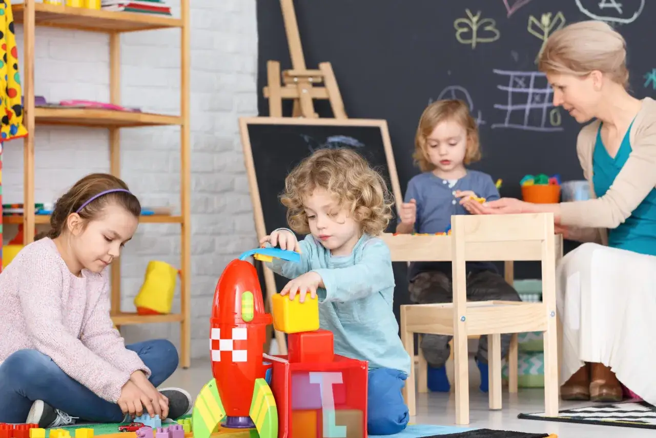 Dzieci bawią się klockami w żłobku, a opiekunka czuwa nad ich bezpieczeństwem. Zastanawiasz się, ile zarabiają opiekunki w żłobku?