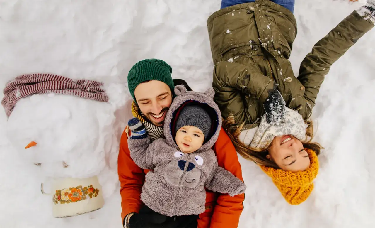 Rodzina cieszy się zimowym dniem. Tata z niemowlakiem w kombinezonie misia, mama leży obok. Idealny czas na spacer, ale jak długo spacerować z niemowlakiem zimą?