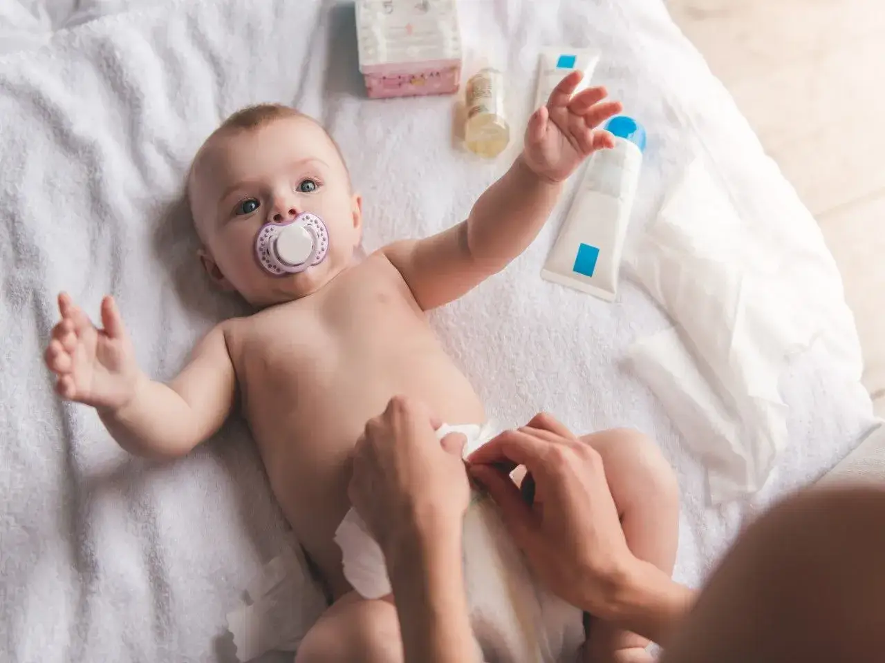 Niemowlę z smoczkiem leży na ręczniku, a ręce rodzica zmieniają mu pieluszkę. Kiedy zmieniać pieluszkę przed czy po karmieniu?