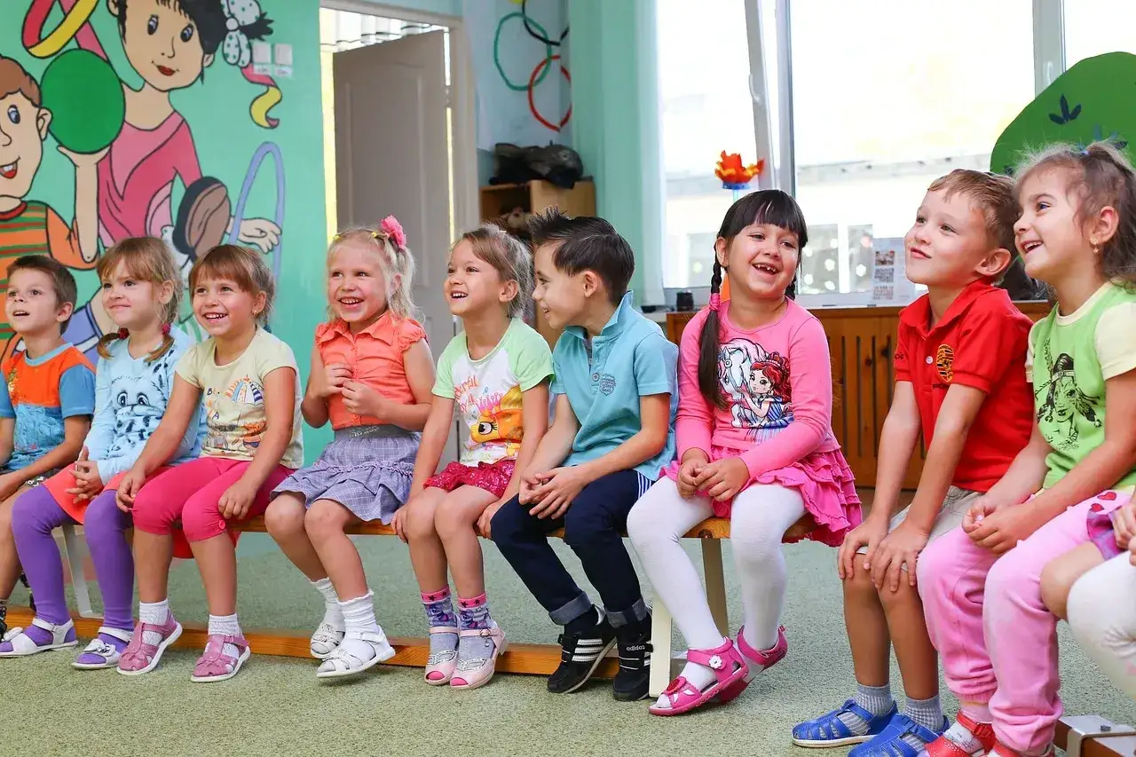 Dzieci w przedszkolu, w wieku od 4 do 6 lat, siedzą na ławce, uśmiechając się i patrząc w górę.