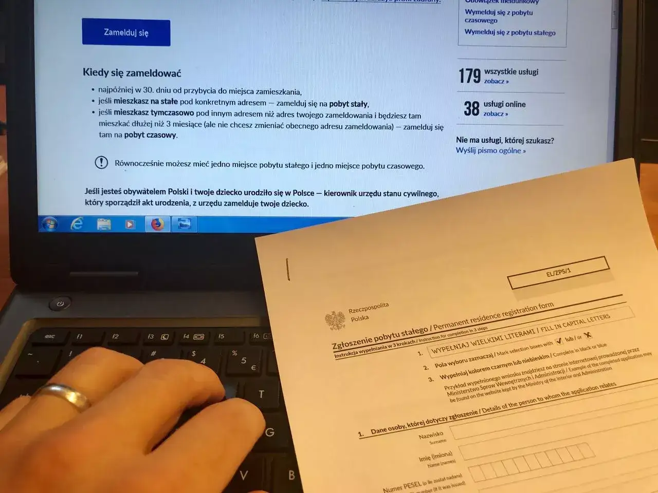 Ręce wpisują dane na formularzu zgłoszenia pobytu stałego, obok ekranu laptopa z informacjami, jak przemeldować dzieci.