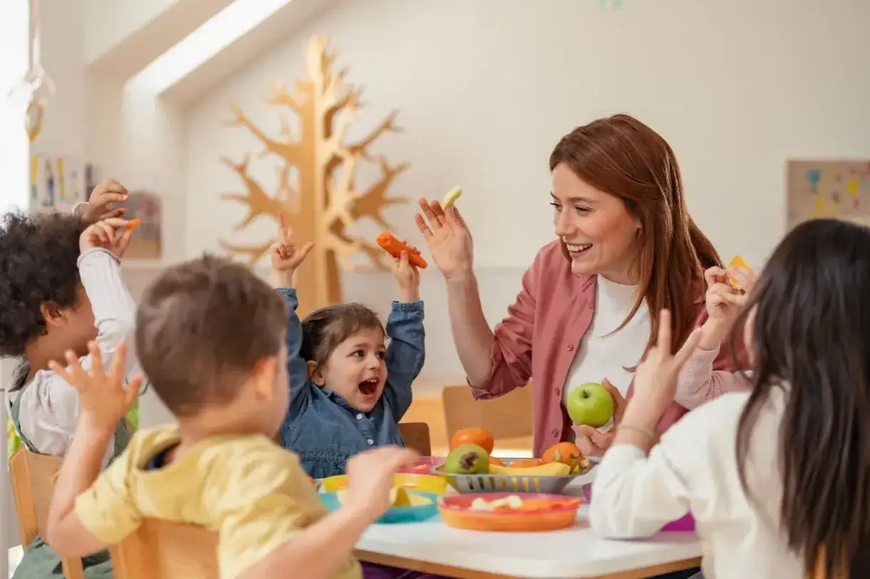 Uśmiechnięta przedszkolanka z dziećmi podczas zajęć o zdrowym jedzeniu. Ile godzin pracuje przedszkolanka? Tyle, ile potrzeba, by widzieć takie radosne buzie!