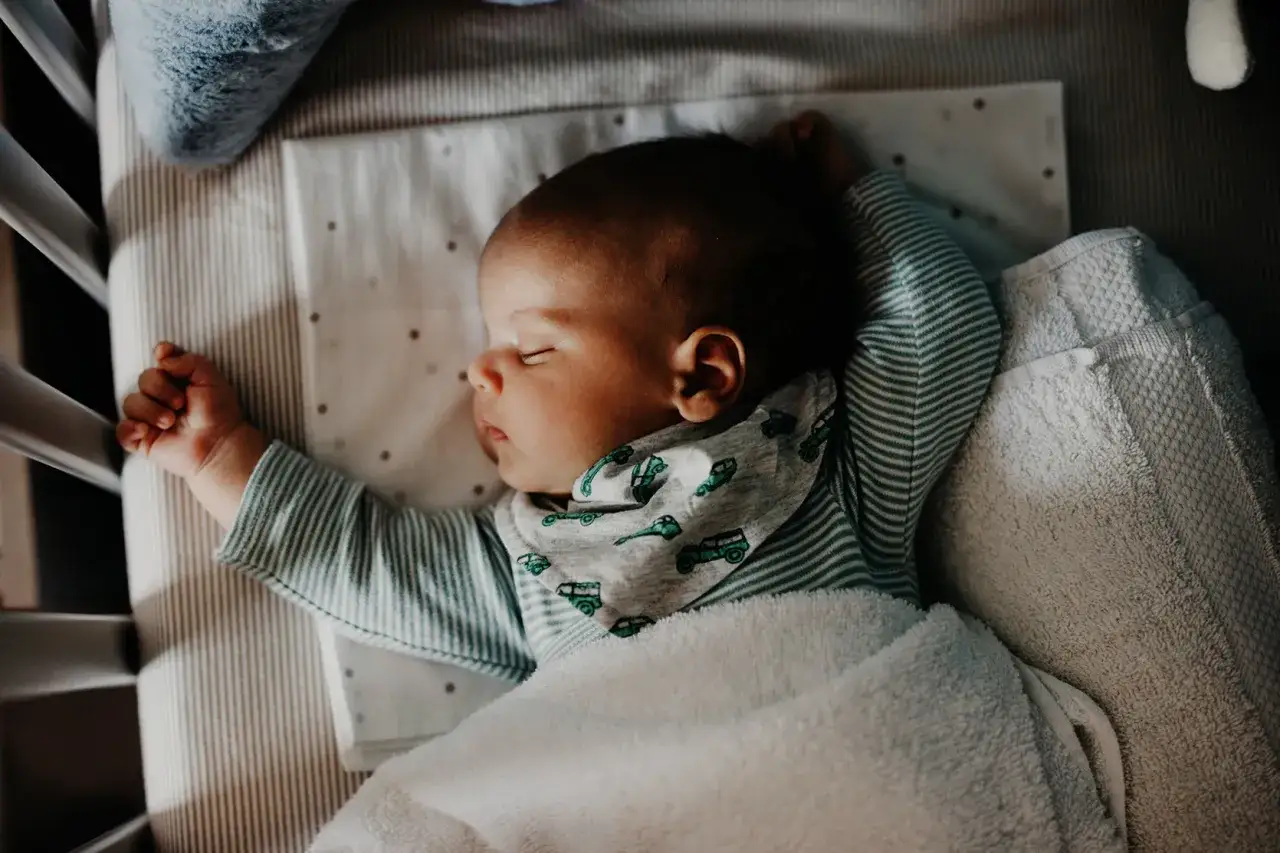 Śpiące niemowlę w łóżeczku, które pokazuje, ile śpi miesięczne dziecko. Otulone kocykiem, w body w paski i z chustką z autkami.