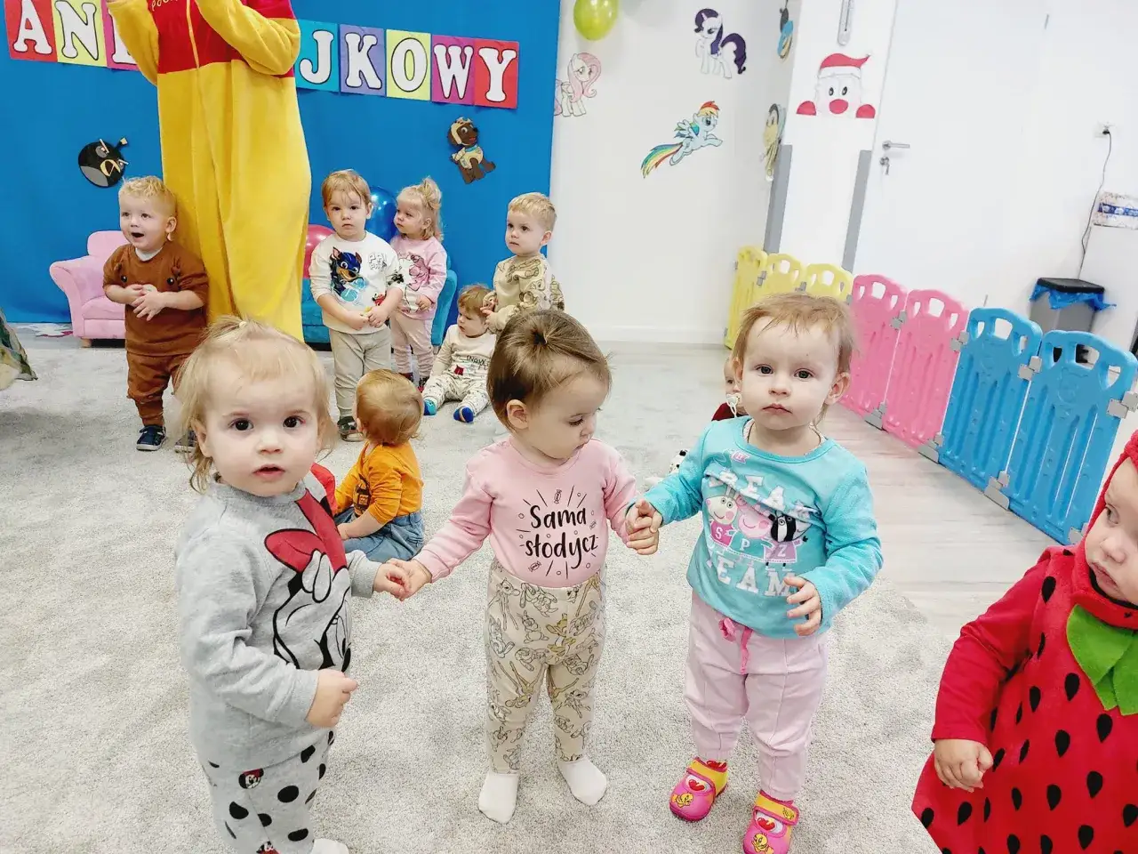 Dzieci trzymają się za ręce w żłobku, zastanawiając się, czy żłobek to dobry pomysł. W tle postać w stroju Kubusia Puchatka.