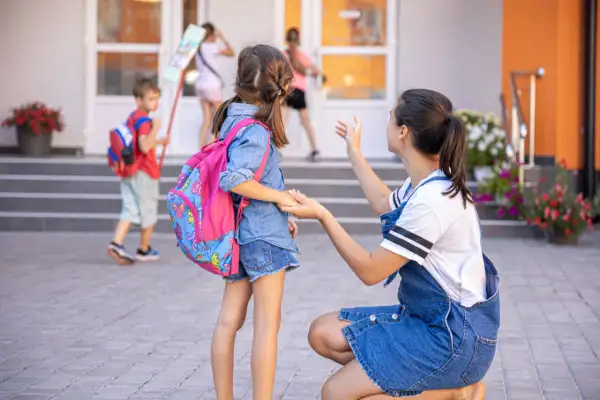 Mama żegna córkę idącą do zerówki. Ile lat ma się w zerówce? Zazwyczaj 6. Dzieci z plecakami wchodzą do szkoły.