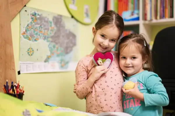 Dwie uśmiechnięte dziewczynki trzymają serduszka. W tle mapa i półki z książkami. Ile godzin w przedszkolu jest bezpłatnych?