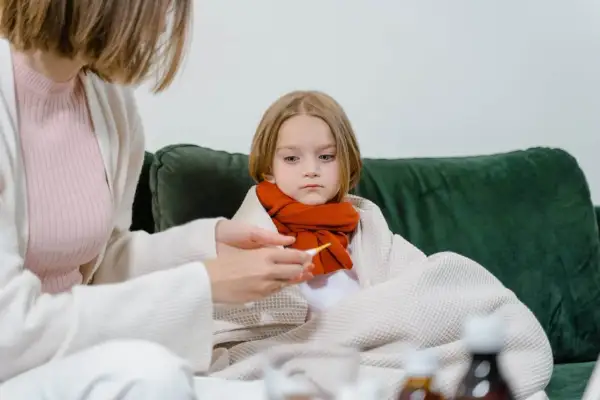 Smutna dziewczynka owinięta kocem, z szalikiem na szyi, czeka na powrót do zdrowia po grypie. Mama mierzy jej temperaturę.
